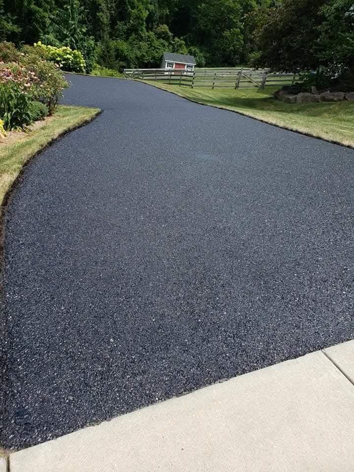 A freshly paved black asphalt driveway curves through a green residential yard toward a small shed in the distance.