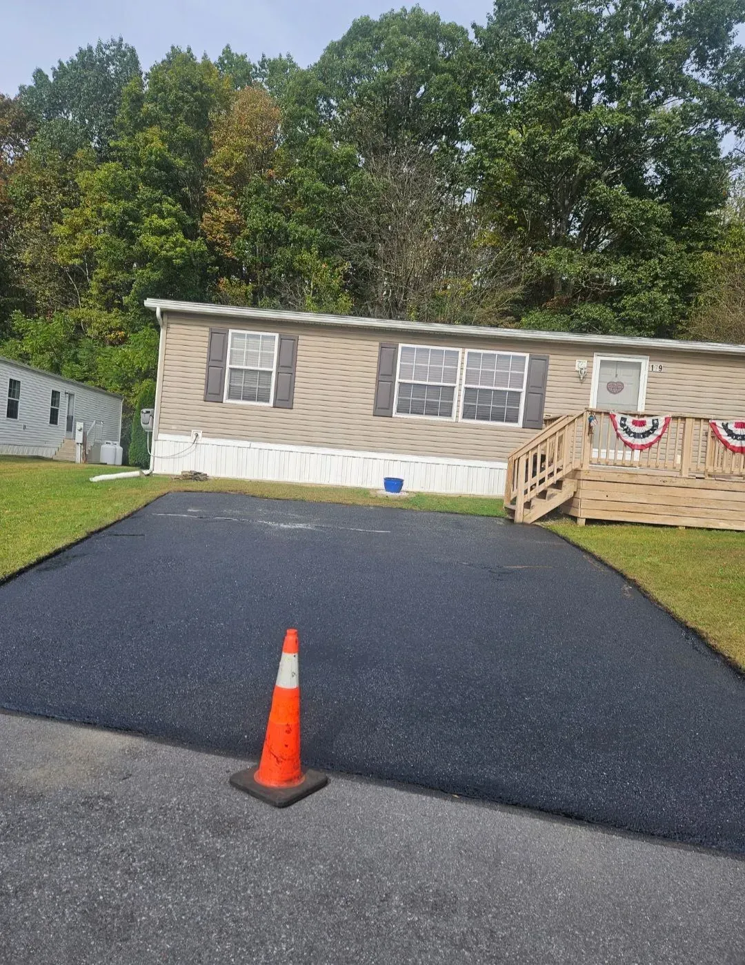 A freshly paved black asphalt driveway sits in front of a tan mobile home with a wooden porch and an orange traffic cone.