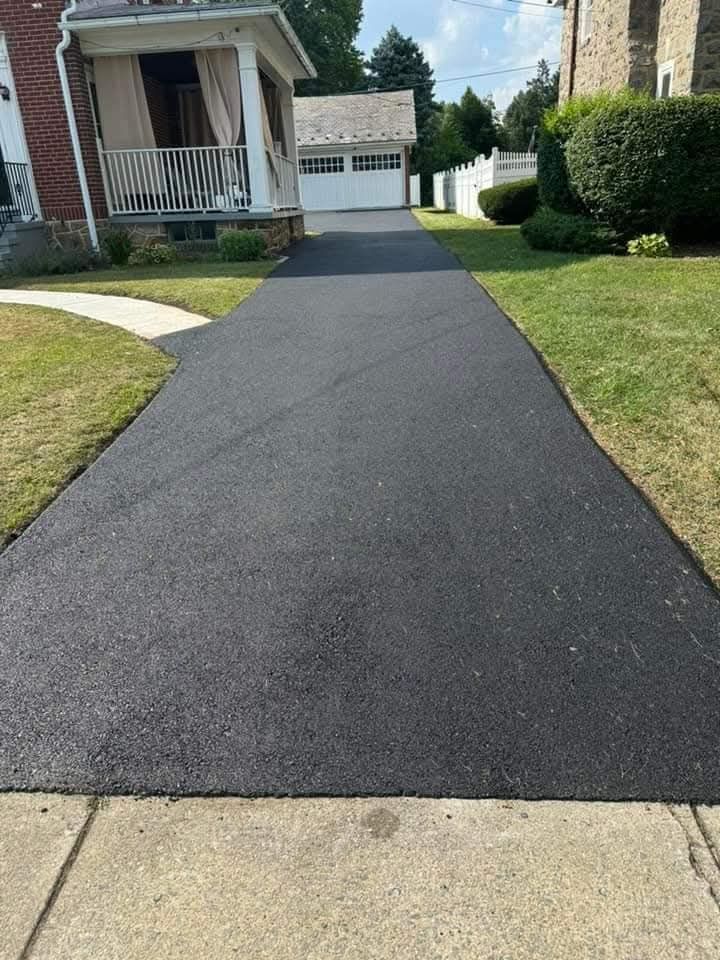A freshly paved, dark asphalt driveway leading toward a white house with a porch and a detached garage in the background.