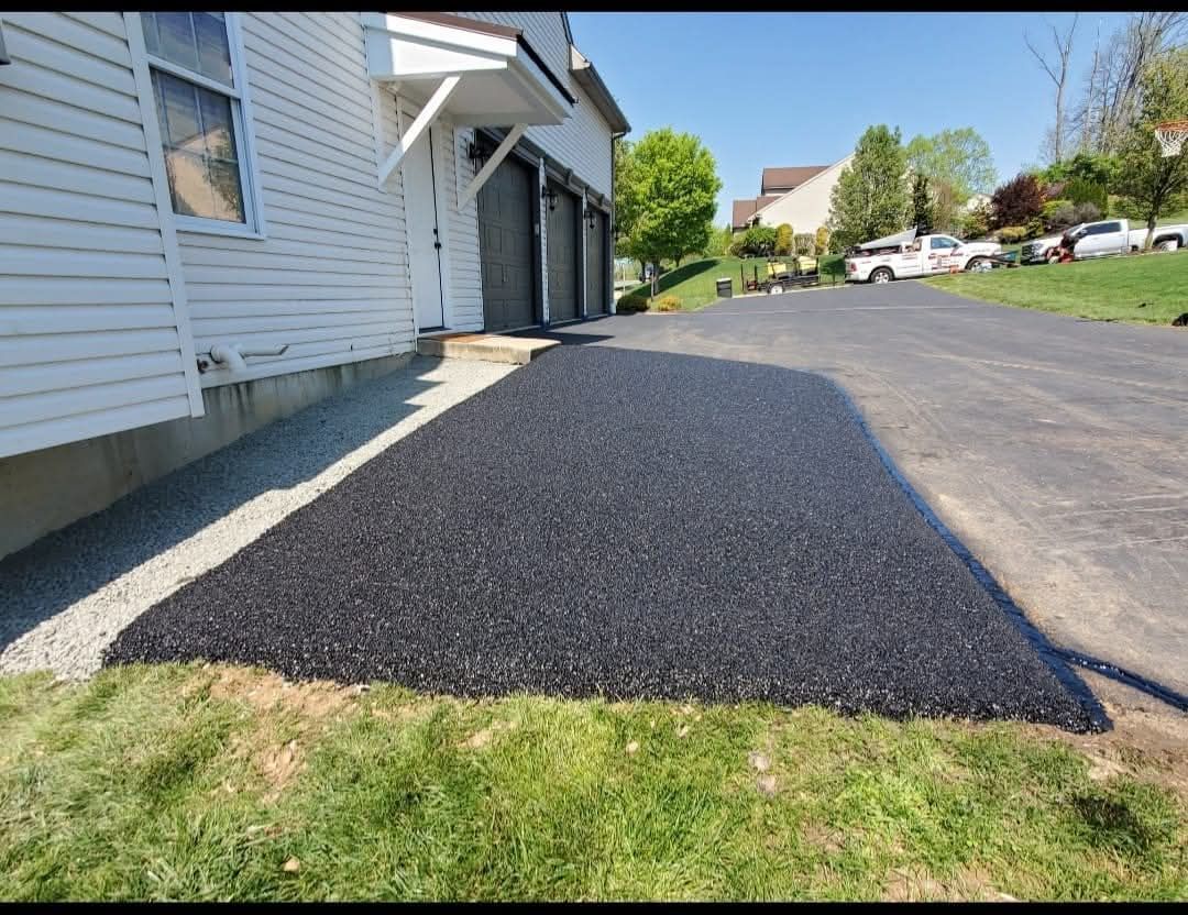 Freshly paved asphalt driveway extension next to a white house with a garage.