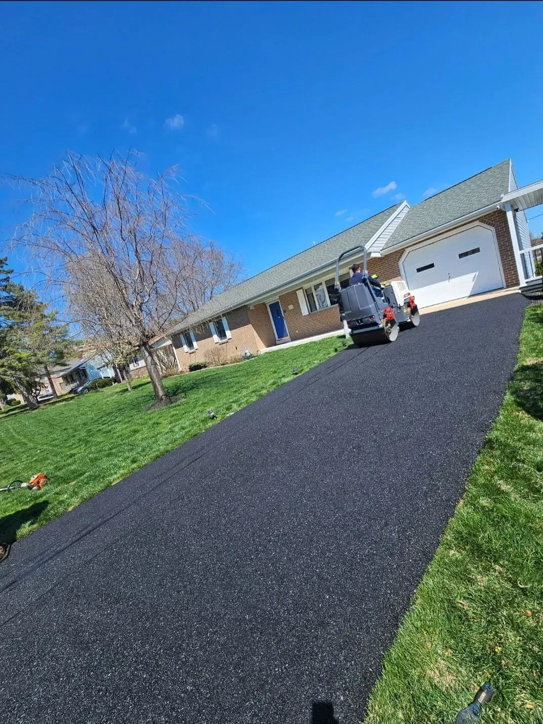 A person operates a steamroller on a newly paved black asphalt driveway leading to a suburban brick house.