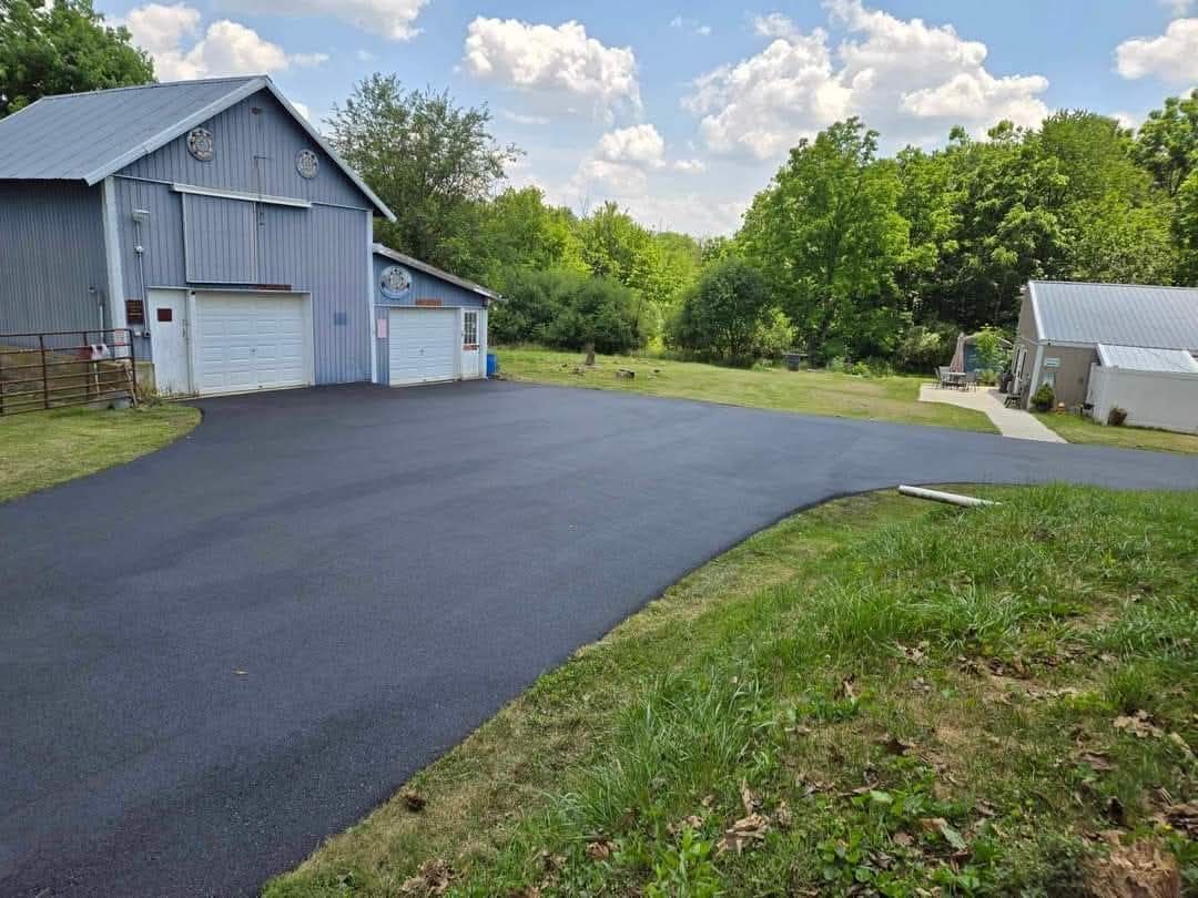 A freshly paved black asphalt driveway leads to a gray barn and a detached garage on a sunny day with green surroundings.