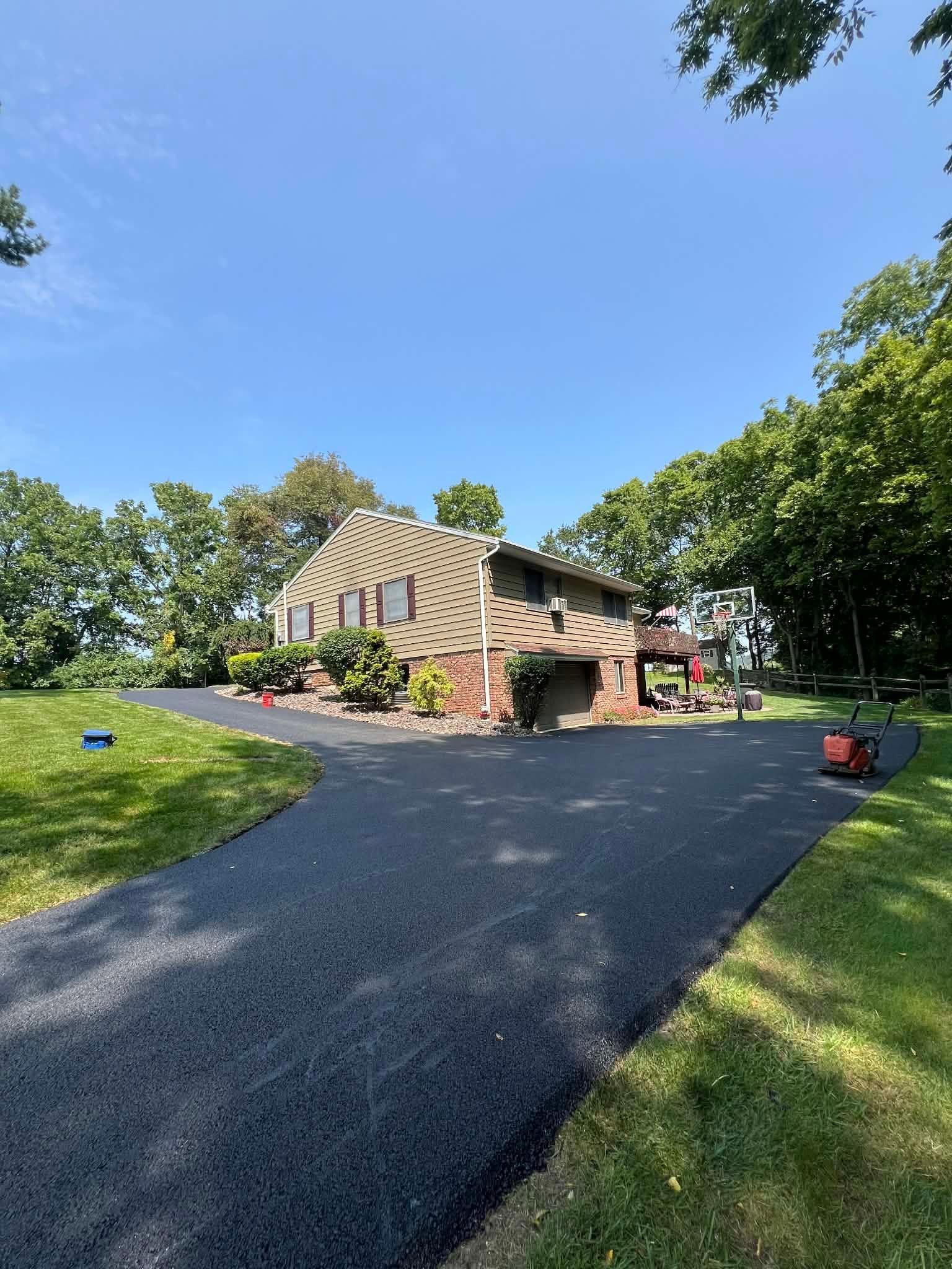 A newly paved asphalt driveway leads up to a split-level house surrounded by green trees on a sunny day.