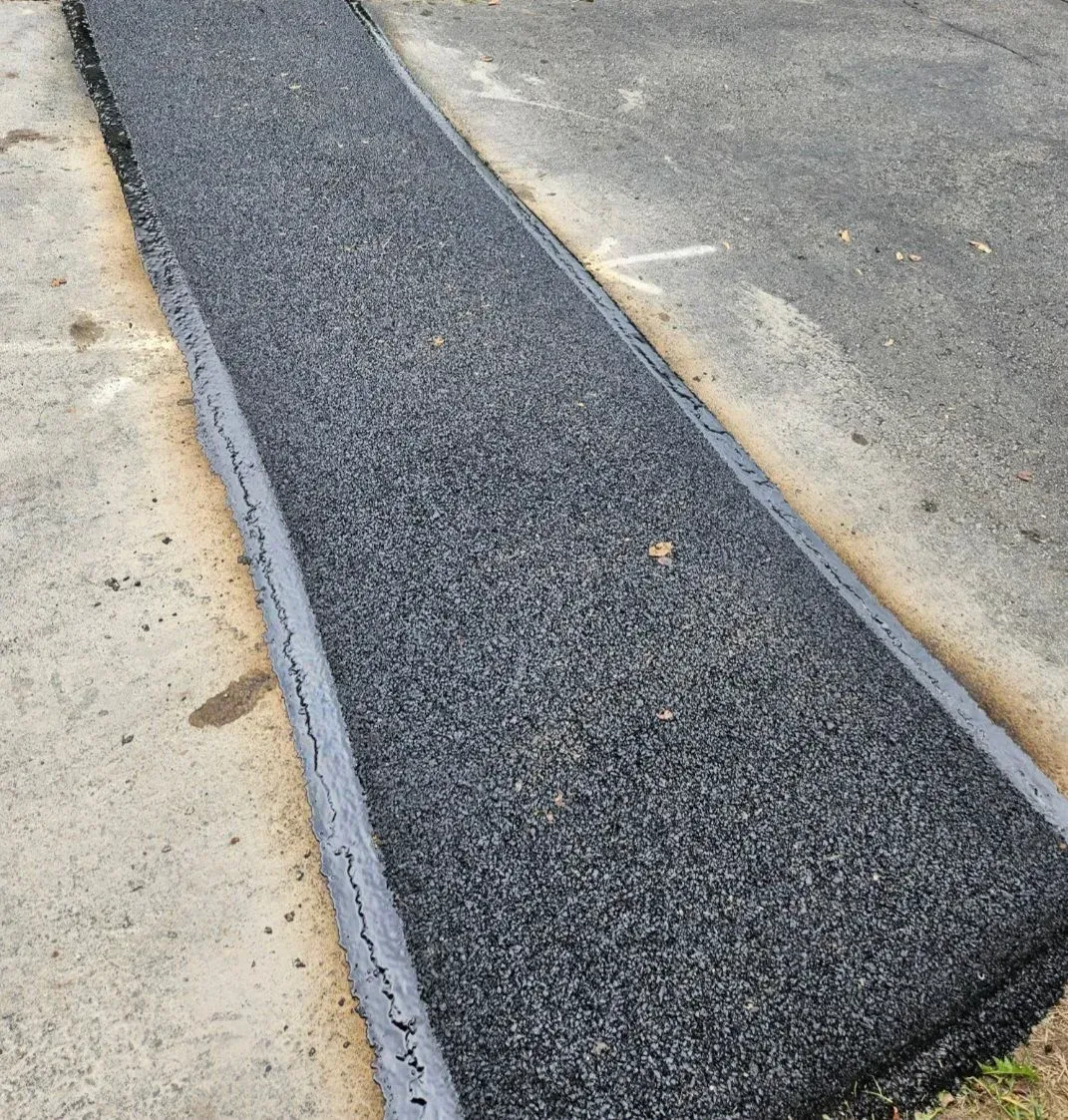 A long strip of newly laid dark asphalt surrounded by a gray concrete border on an outdoor walkway.