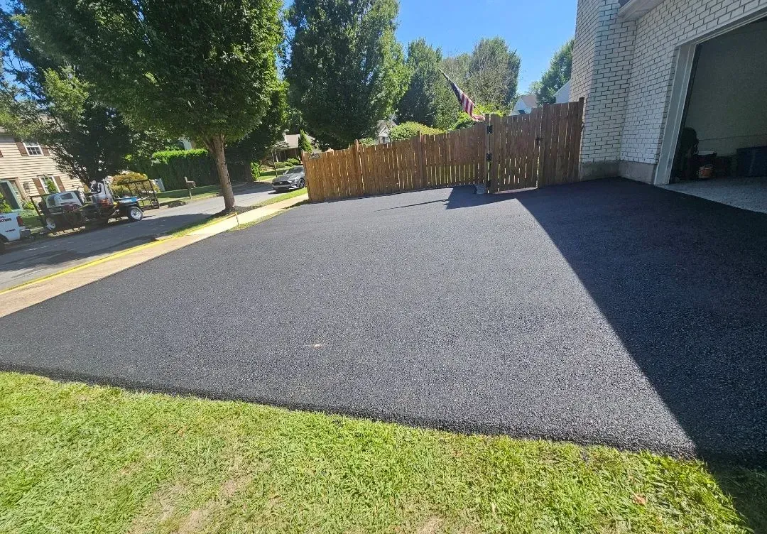 A newly paved black asphalt driveway leading to a garage next to a wooden fence and grassy lawn on a sunny day.