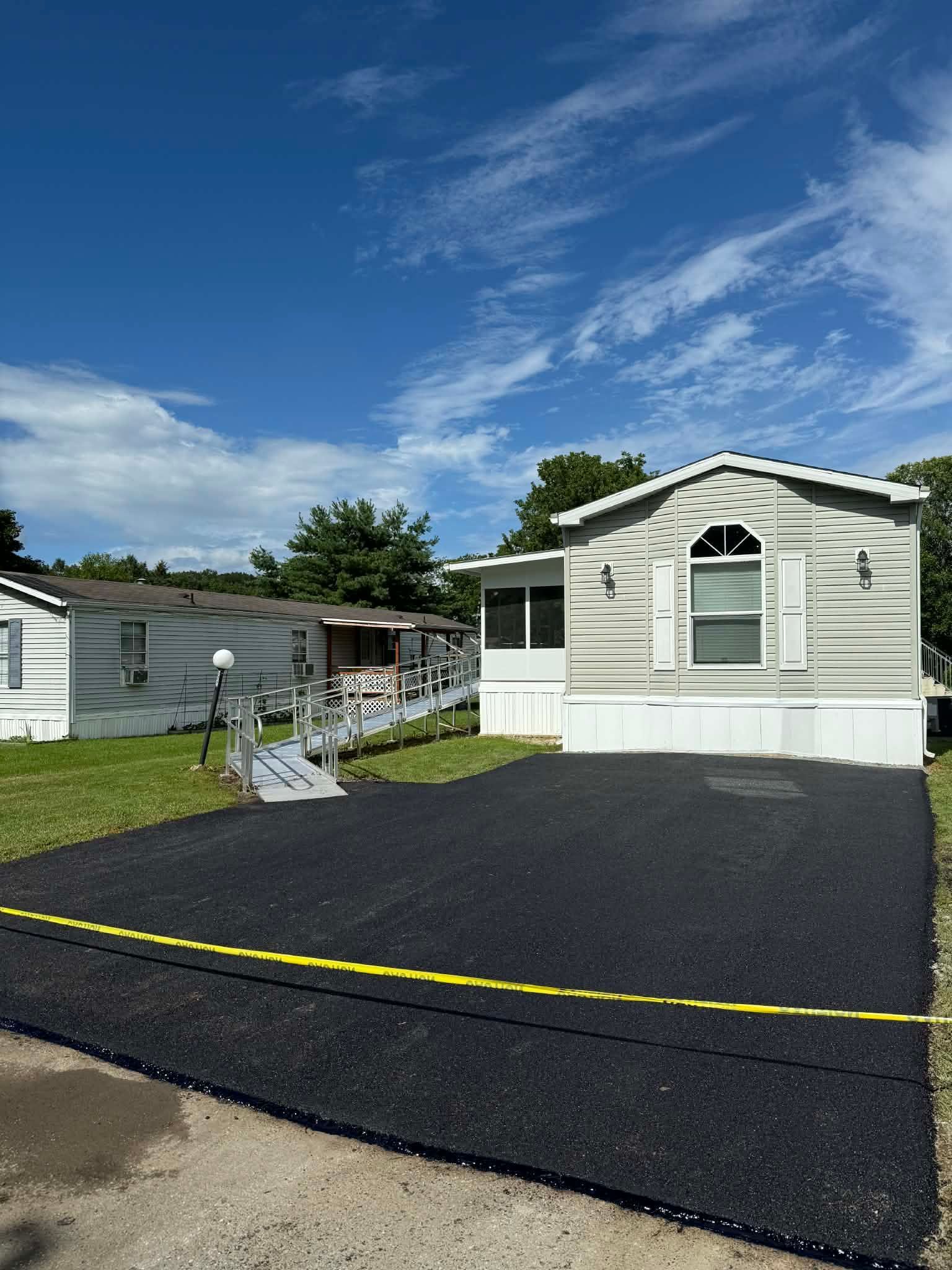 A fresh black asphalt driveway sits in front of a light-colored mobile home under a clear blue sky.