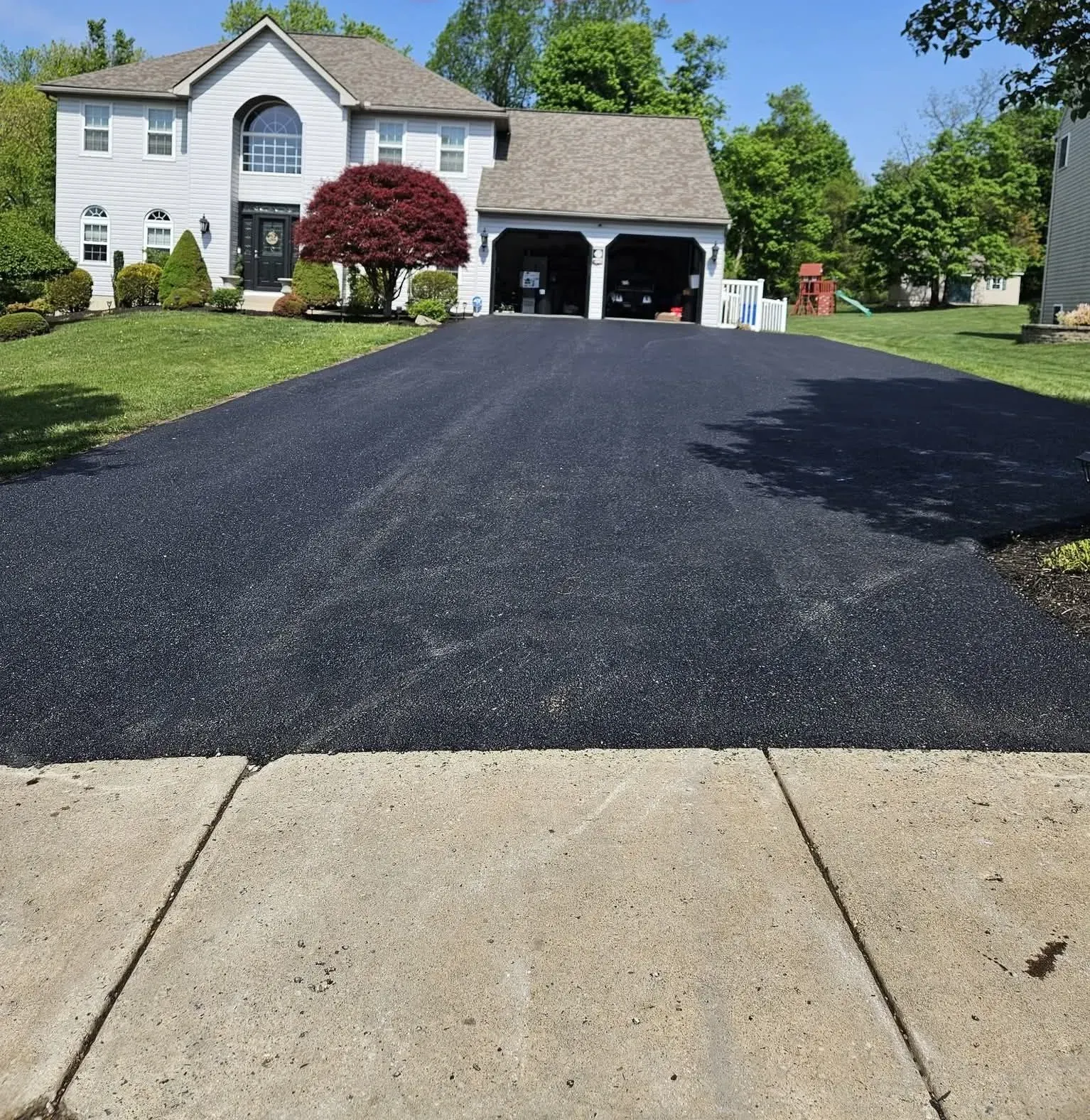 A freshly paved black asphalt driveway leads to a large, two-story house with an attached garage on a sunny day.