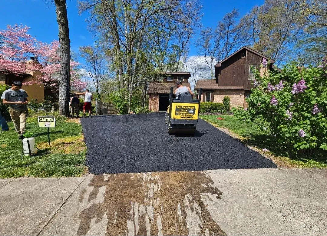 A construction worker operates a yellow roller to smooth fresh black asphalt on a driveway in a residential yard.