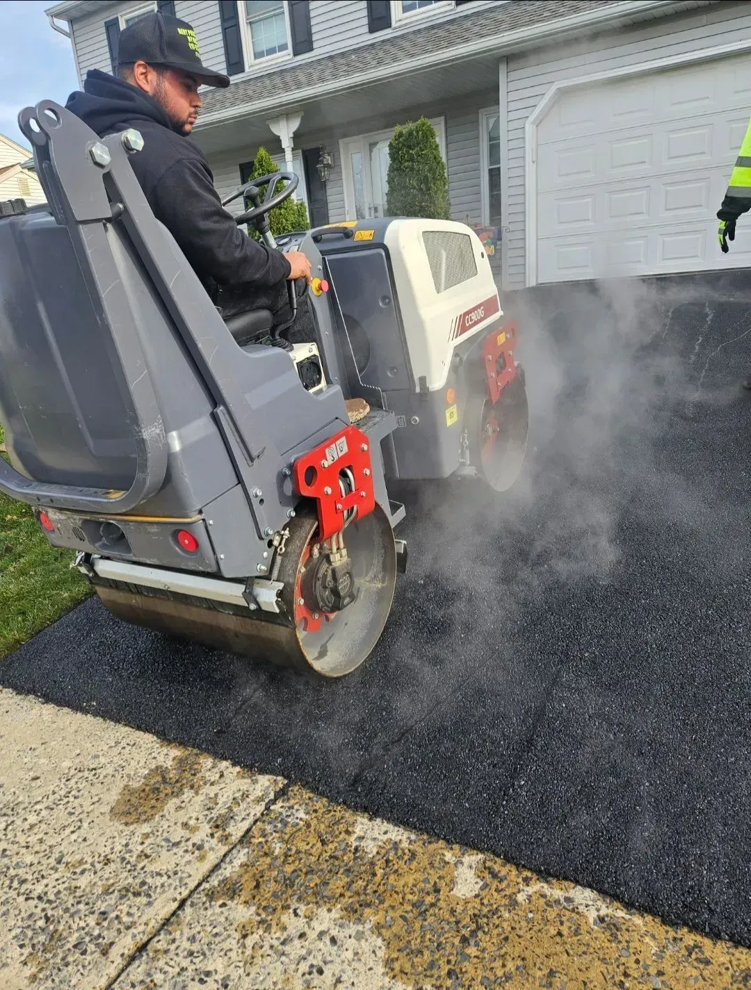 A person operates a steamroller to compact fresh, steaming asphalt on a residential driveway.