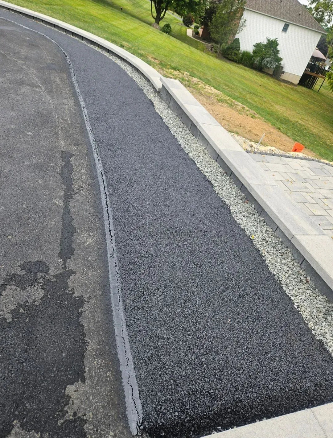 A freshly paved black asphalt strip runs alongside a concrete curb and a gravel border next to a residential lawn.