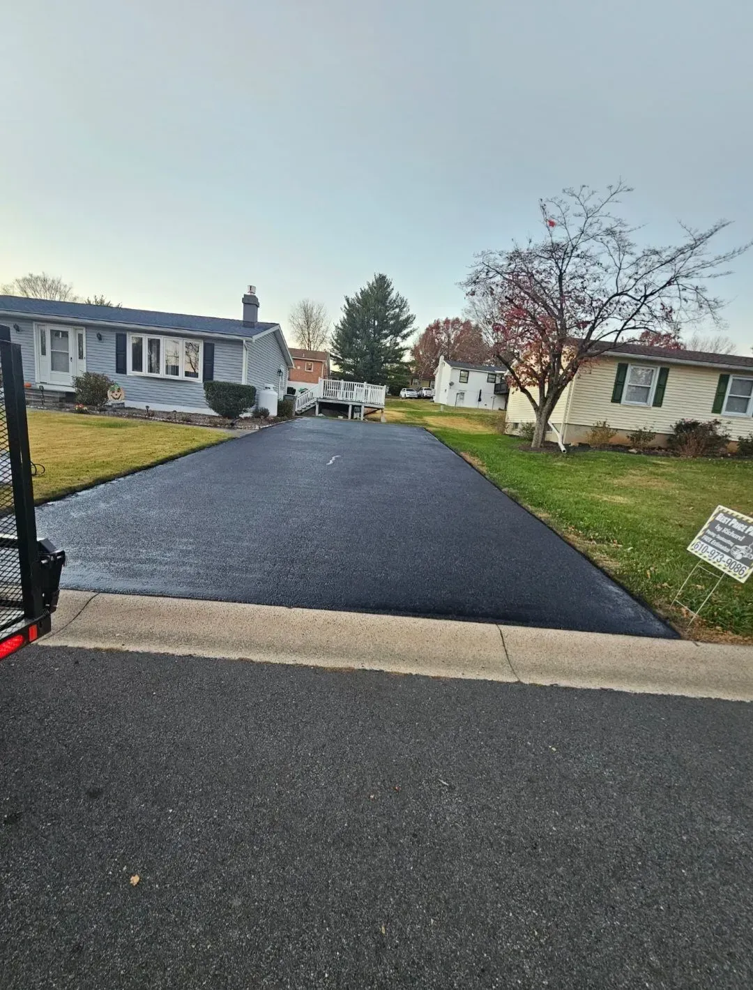 A freshly paved black asphalt driveway between two houses in a residential neighborhood under a clear sky.