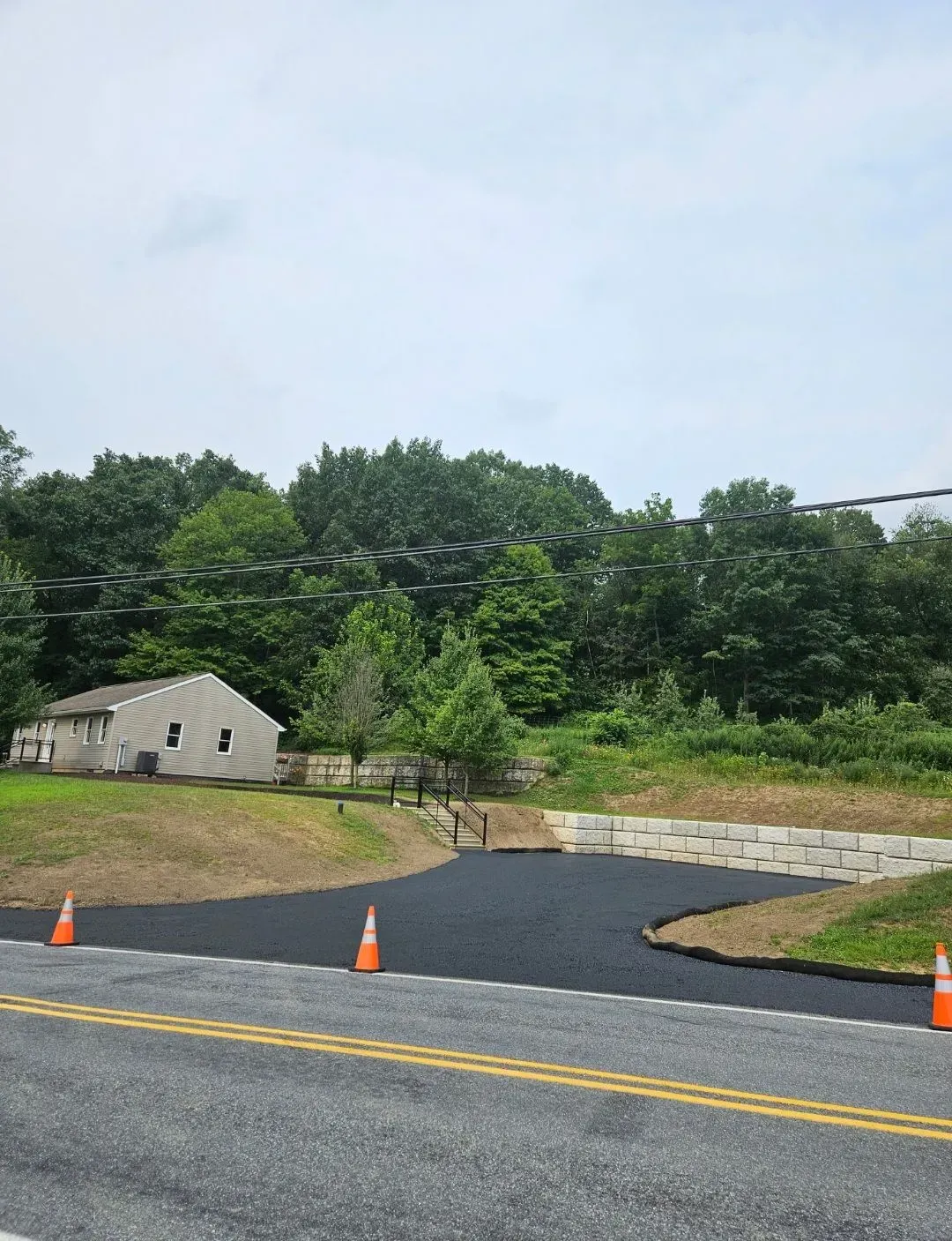 A freshly paved asphalt driveway meets a paved road, bordered by a concrete retaining wall, grassy slopes, and trees.