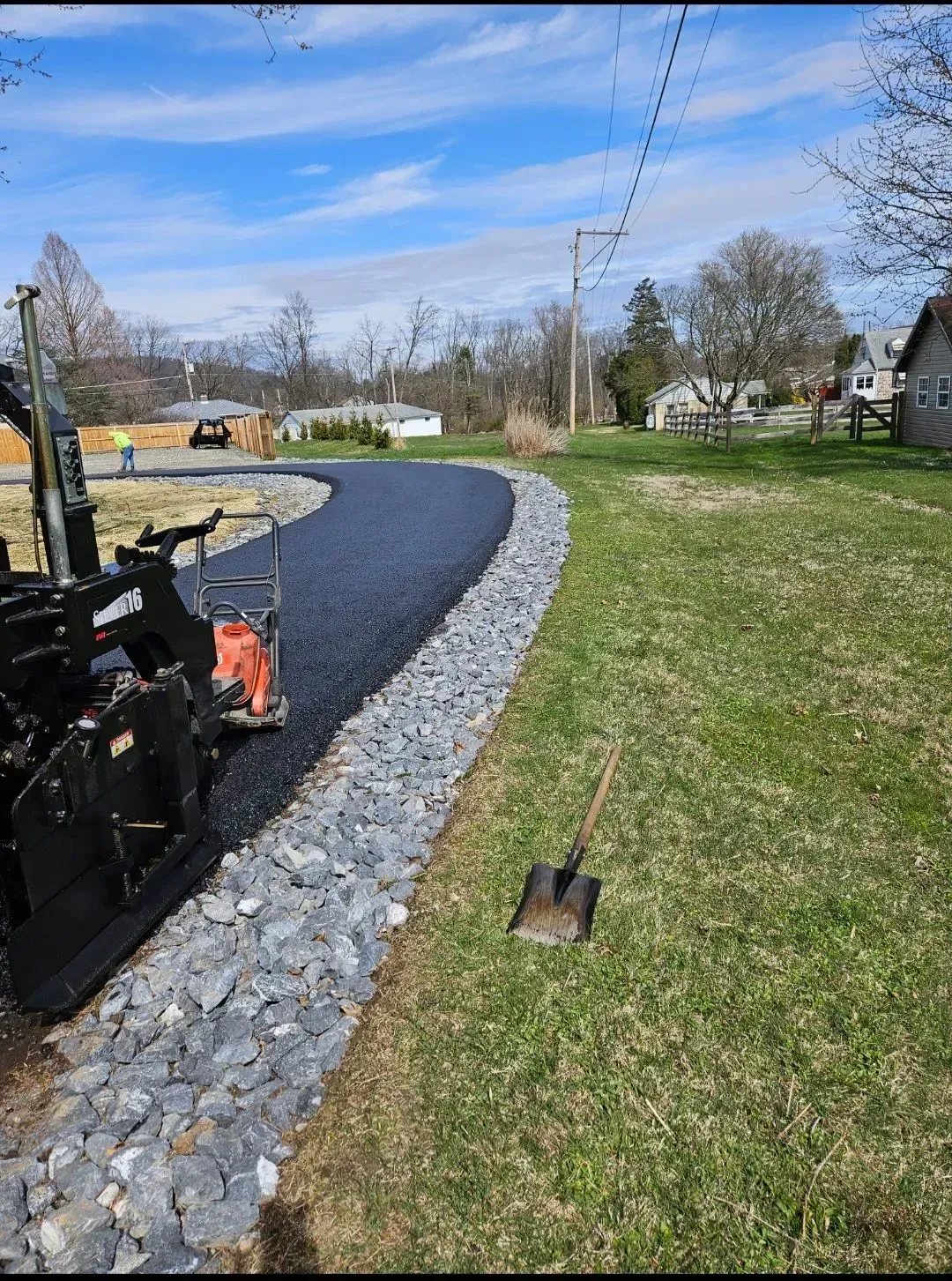 A freshly paved driveway curves through a grassy yard, bordered by large grey rocks with a shovel resting nearby.