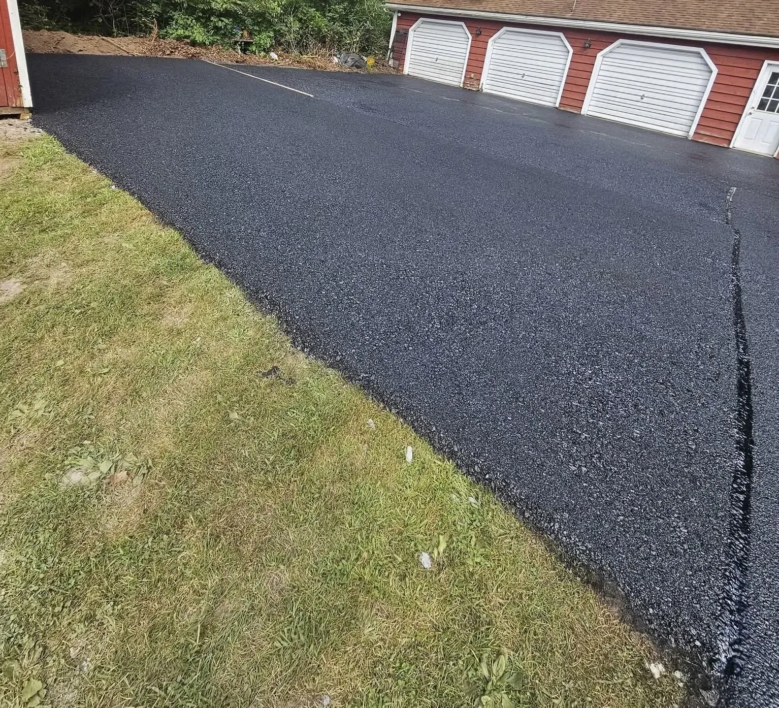 A freshly paved, black asphalt driveway beside a patch of green grass and a house with three white garage doors.
