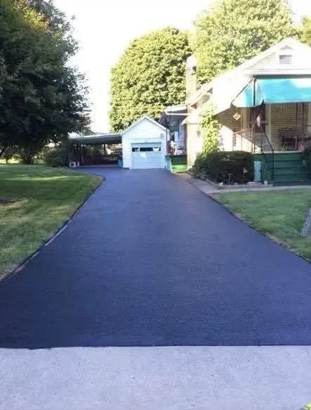 A freshly paved, dark asphalt driveway leads toward a detached white garage next to a house with a turquoise awning.