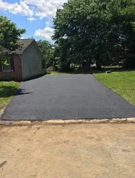 A freshly paved asphalt driveway sits between a small house on the left and a grassy yard with a large tree.