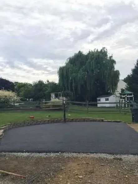 A freshly paved dark asphalt driveway area sits in a grassy backyard with a large willow tree in the background.
