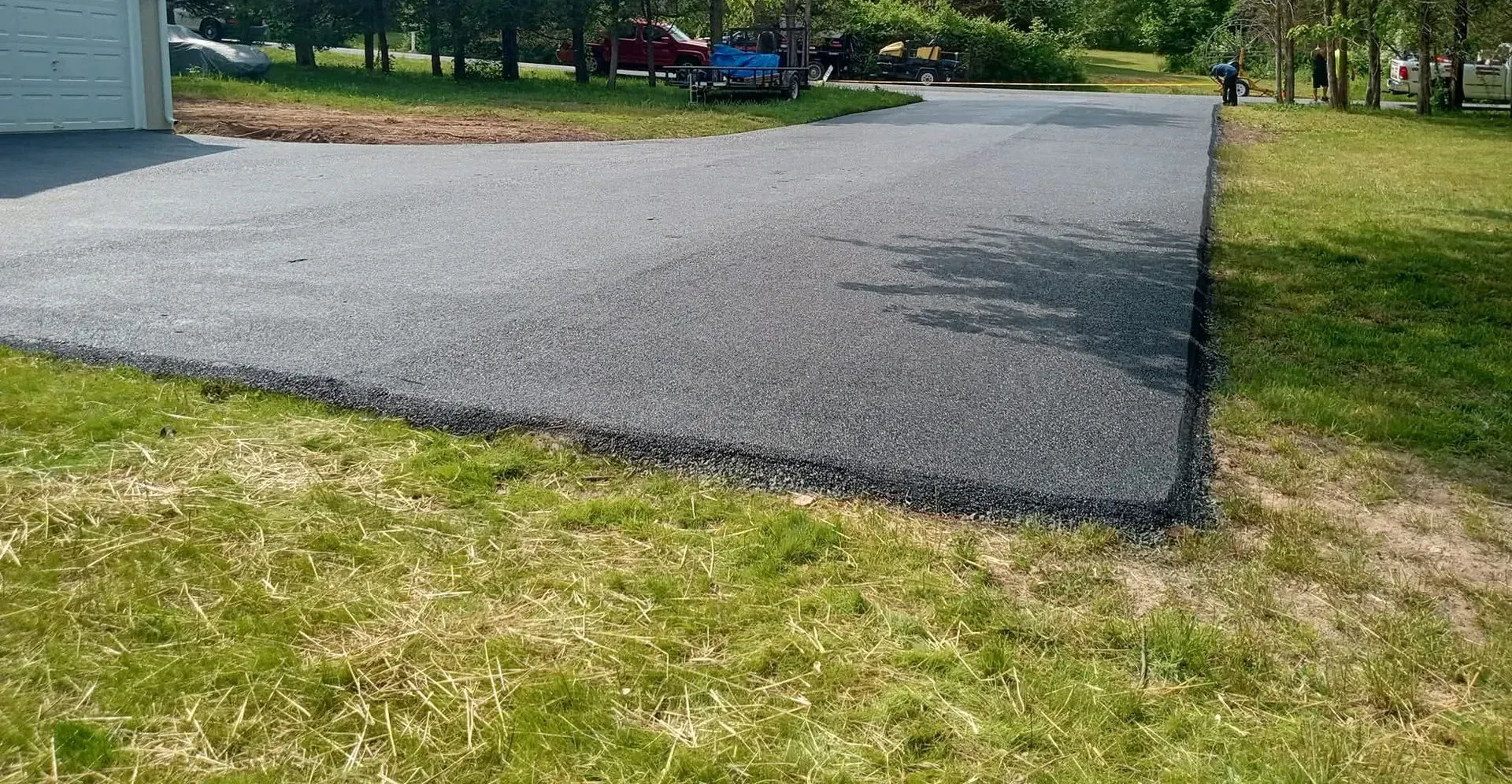 A freshly paved dark asphalt driveway leads to a garage, bordered by green grass and trees in a residential setting.