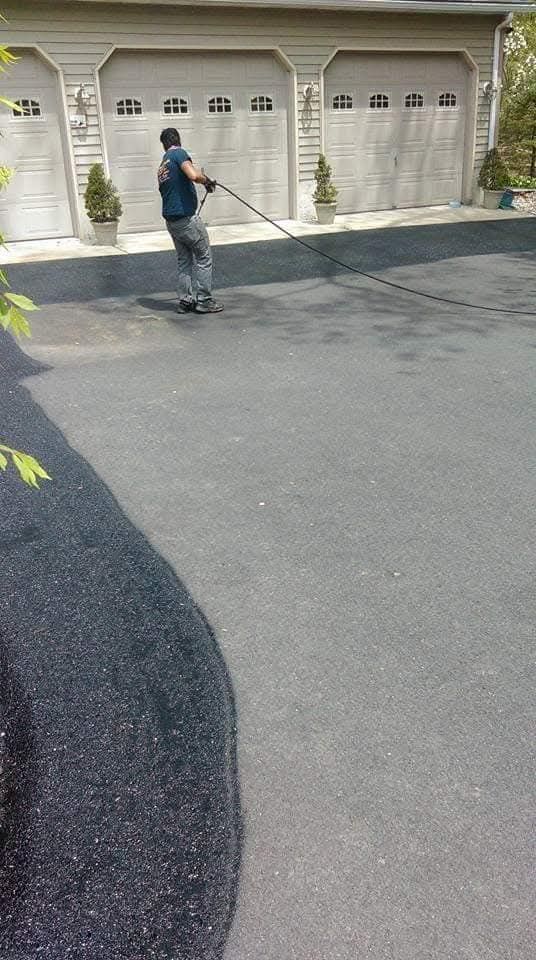 A person in a blue shirt and gray pants pulls a long black hose across a driveway toward a three-car garage.