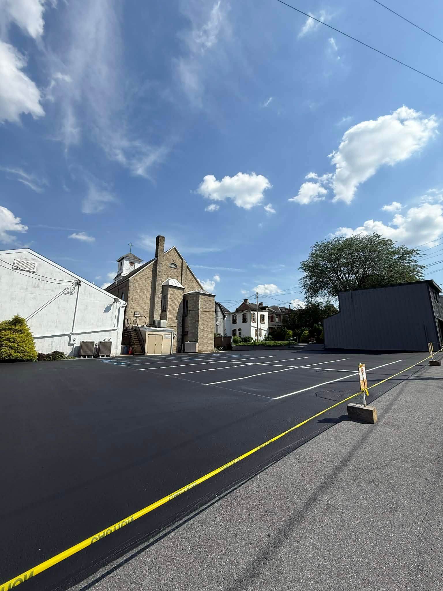 A newly paved parking lot with white-lined spaces and a yellow edge, situated beside a stone building under a blue sky.