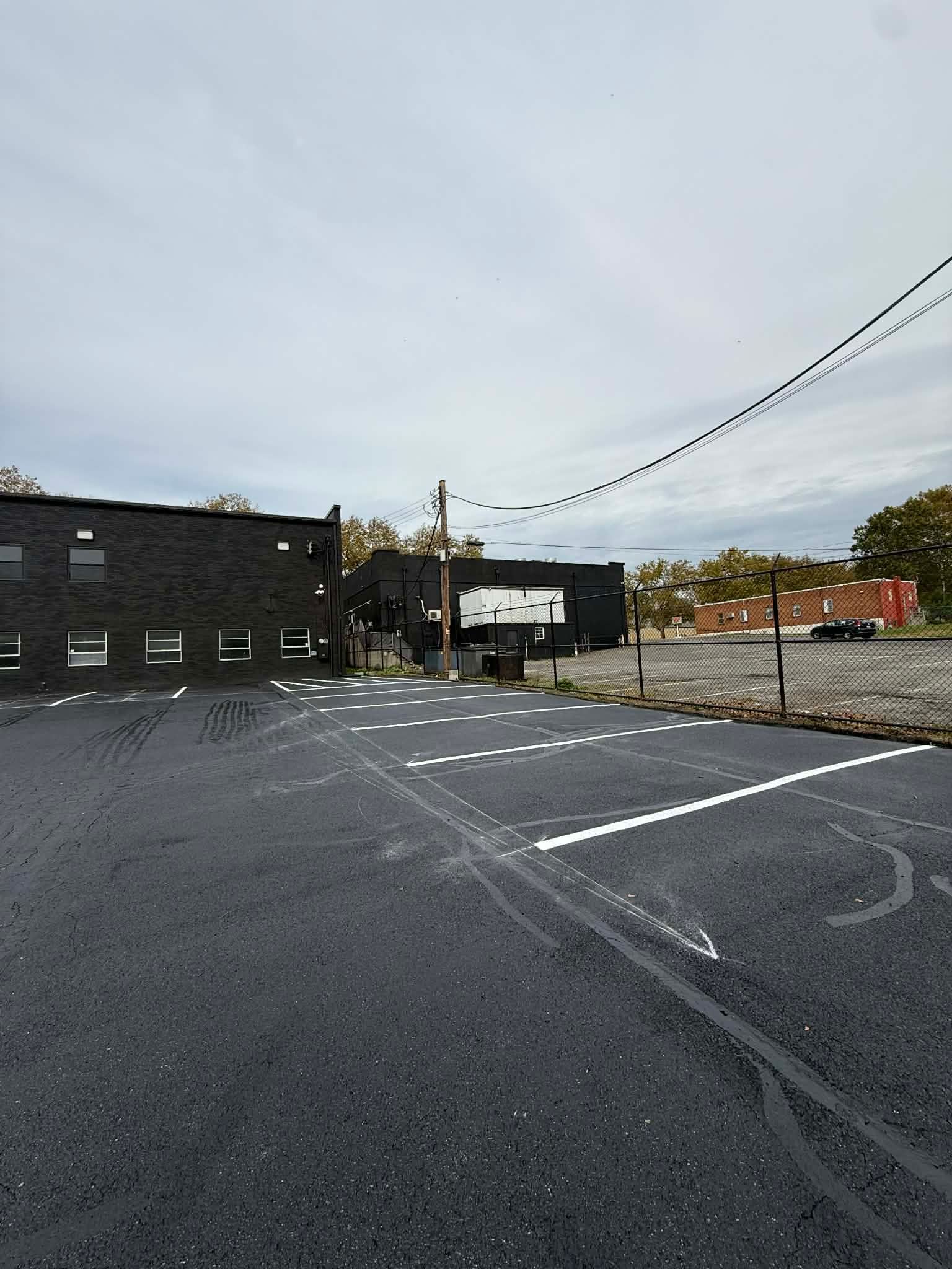 An empty parking lot with painted white stalls, bordering a black brick building under a cloudy, overcast sky.