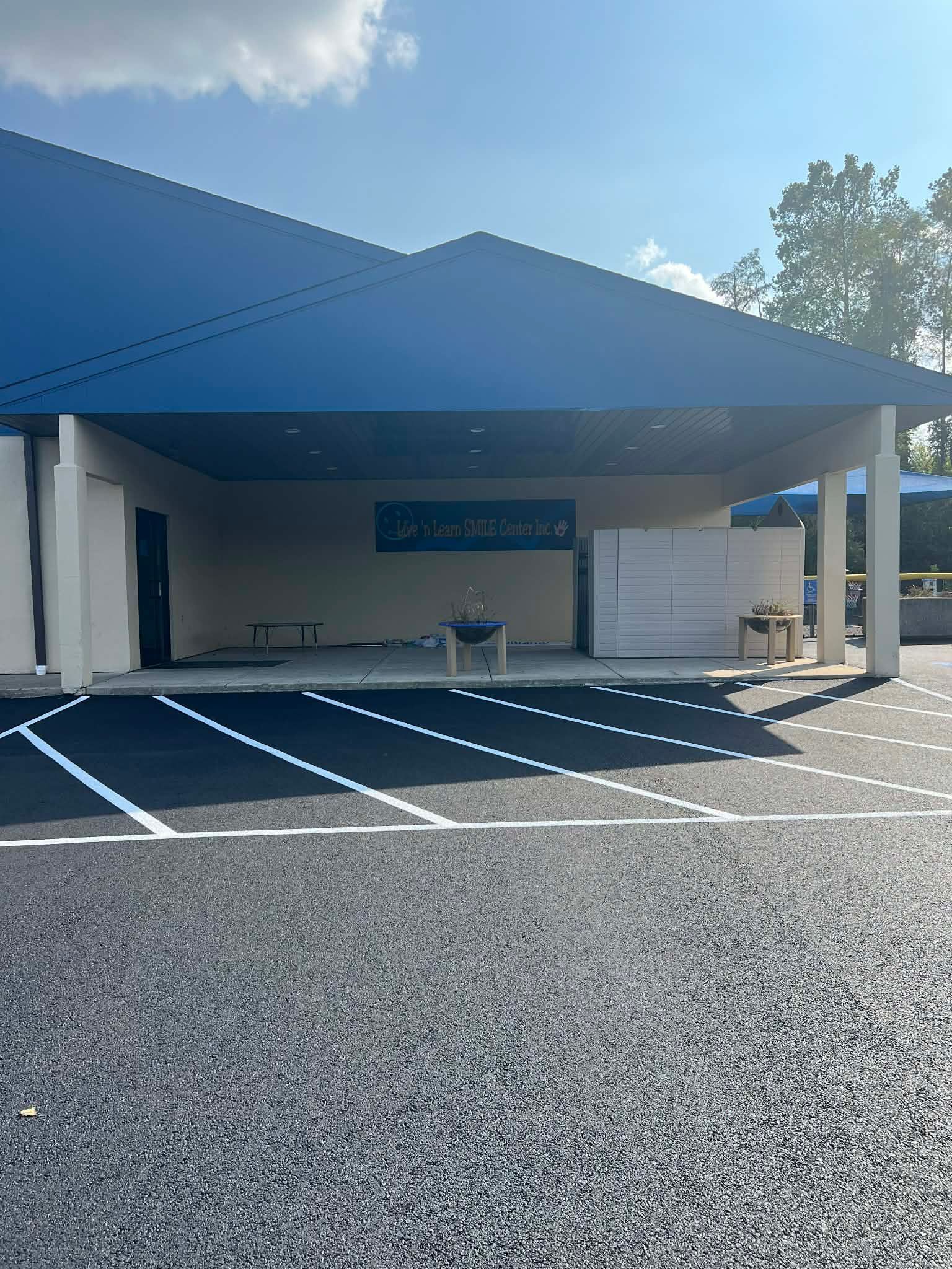 A parking lot leads to a building with a blue roof, a wide shaded entrance, and a small, lone table under the canopy.
