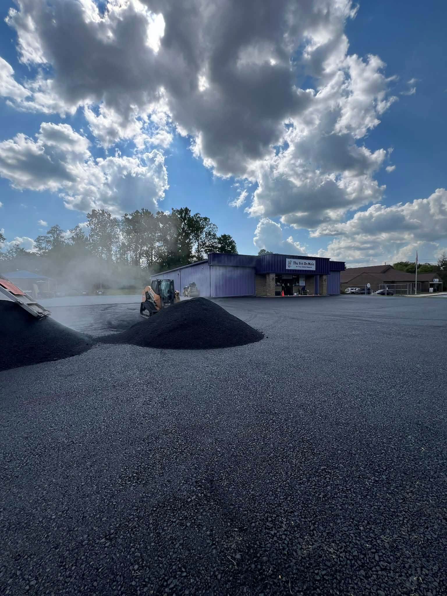 A construction vehicle levels fresh asphalt in a large parking lot under a cloudy blue sky.