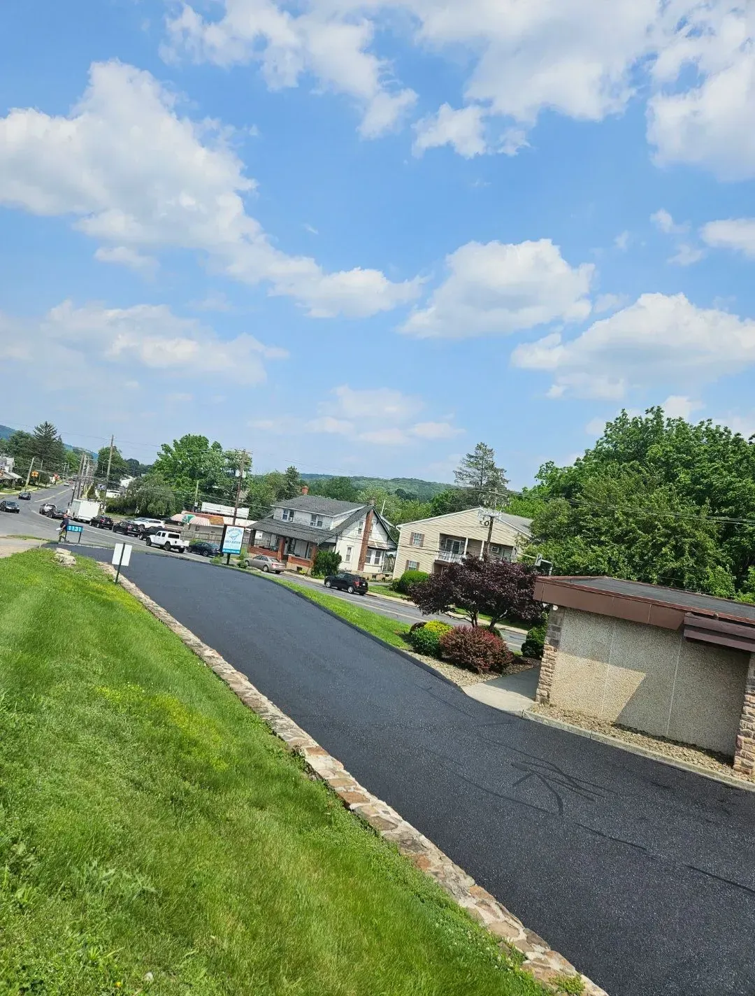 A freshly paved, dark asphalt driveway slopes down toward a street with houses and cars under a blue sky with clouds.