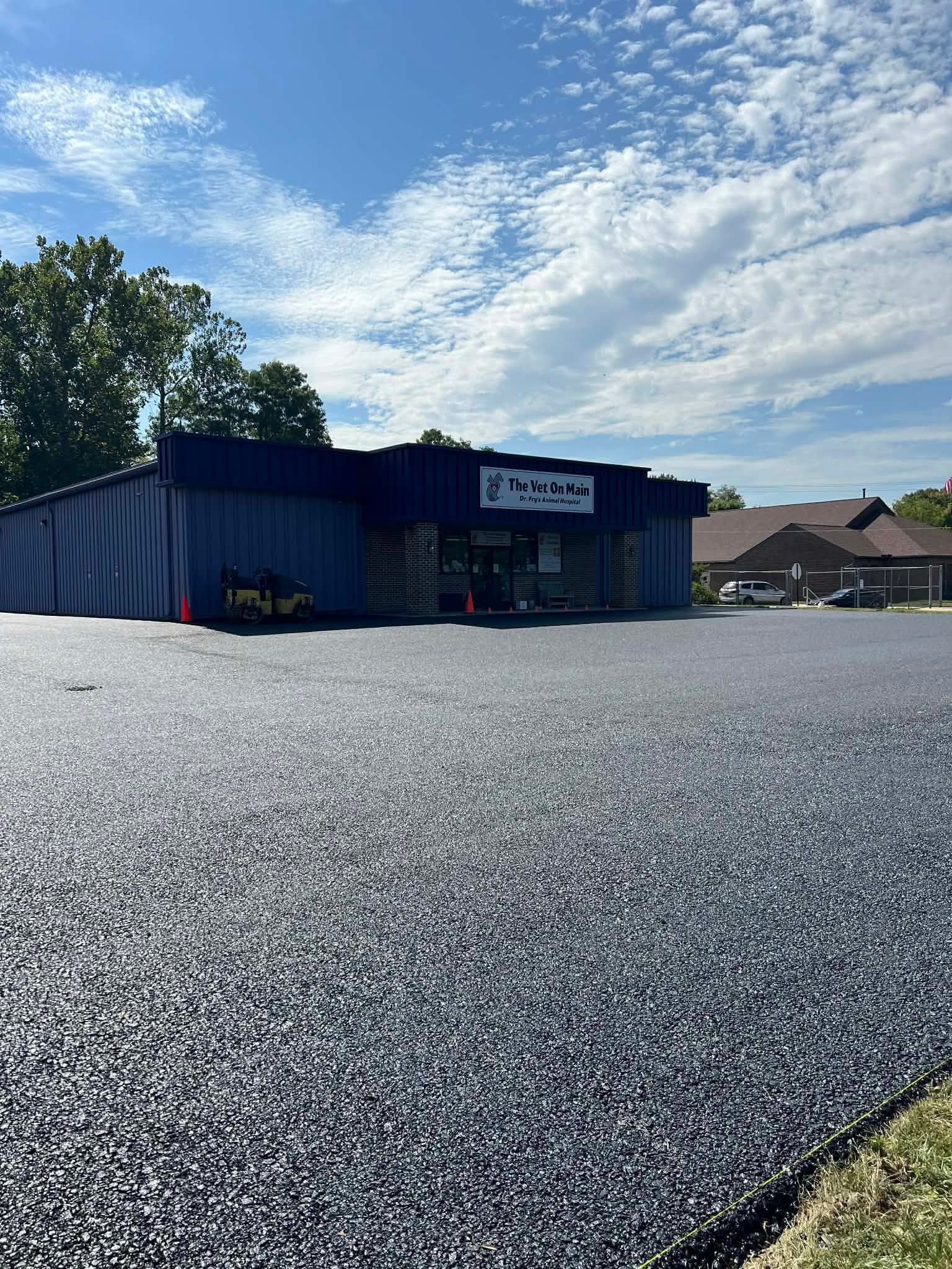 A blue-sided building with a sign sits behind a newly paved, dark asphalt parking lot under a blue, cloudy sky.