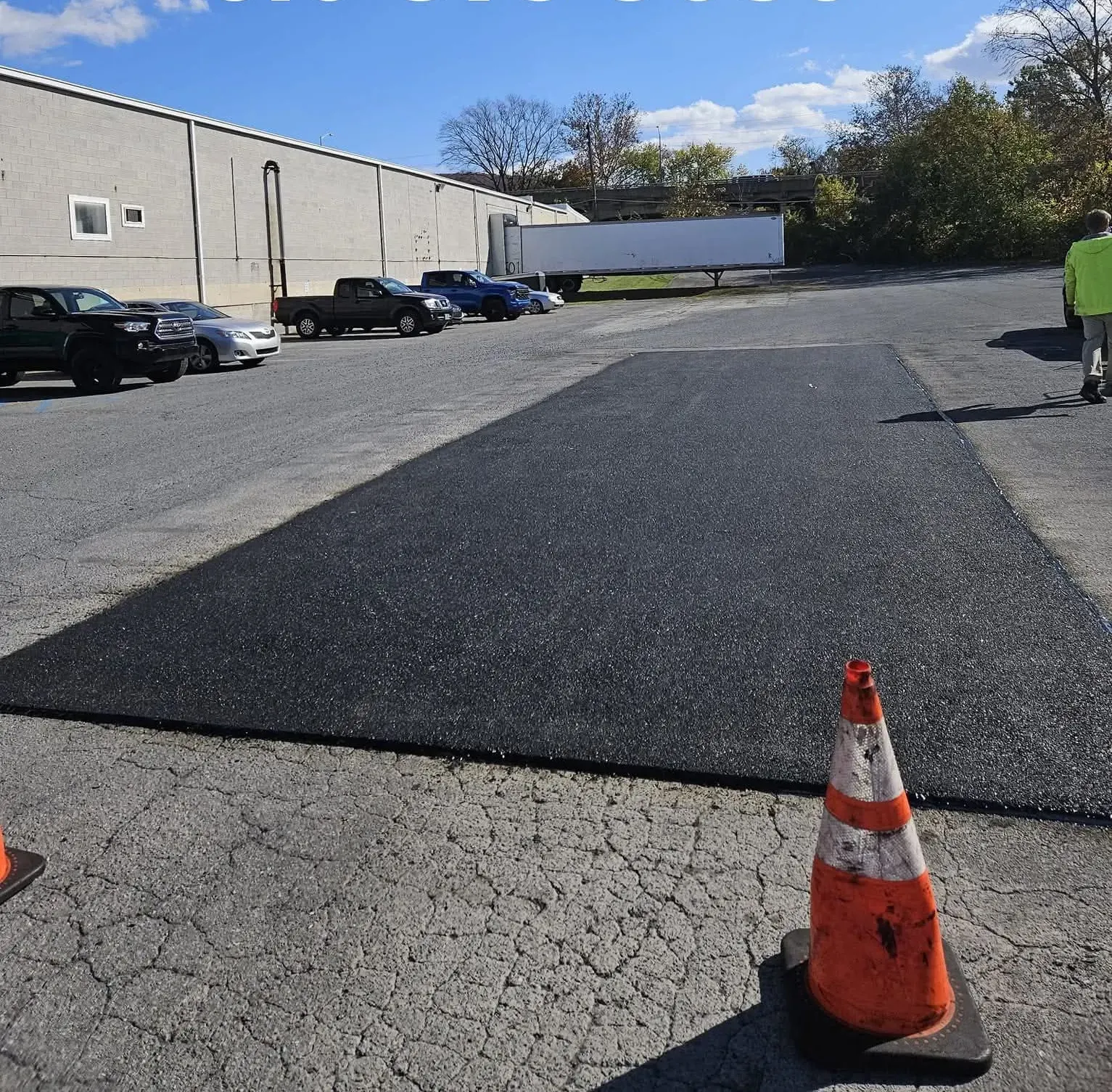 A freshly paved asphalt patch sits in a gravel lot next to a warehouse, marked by an orange traffic cone in the foreground.