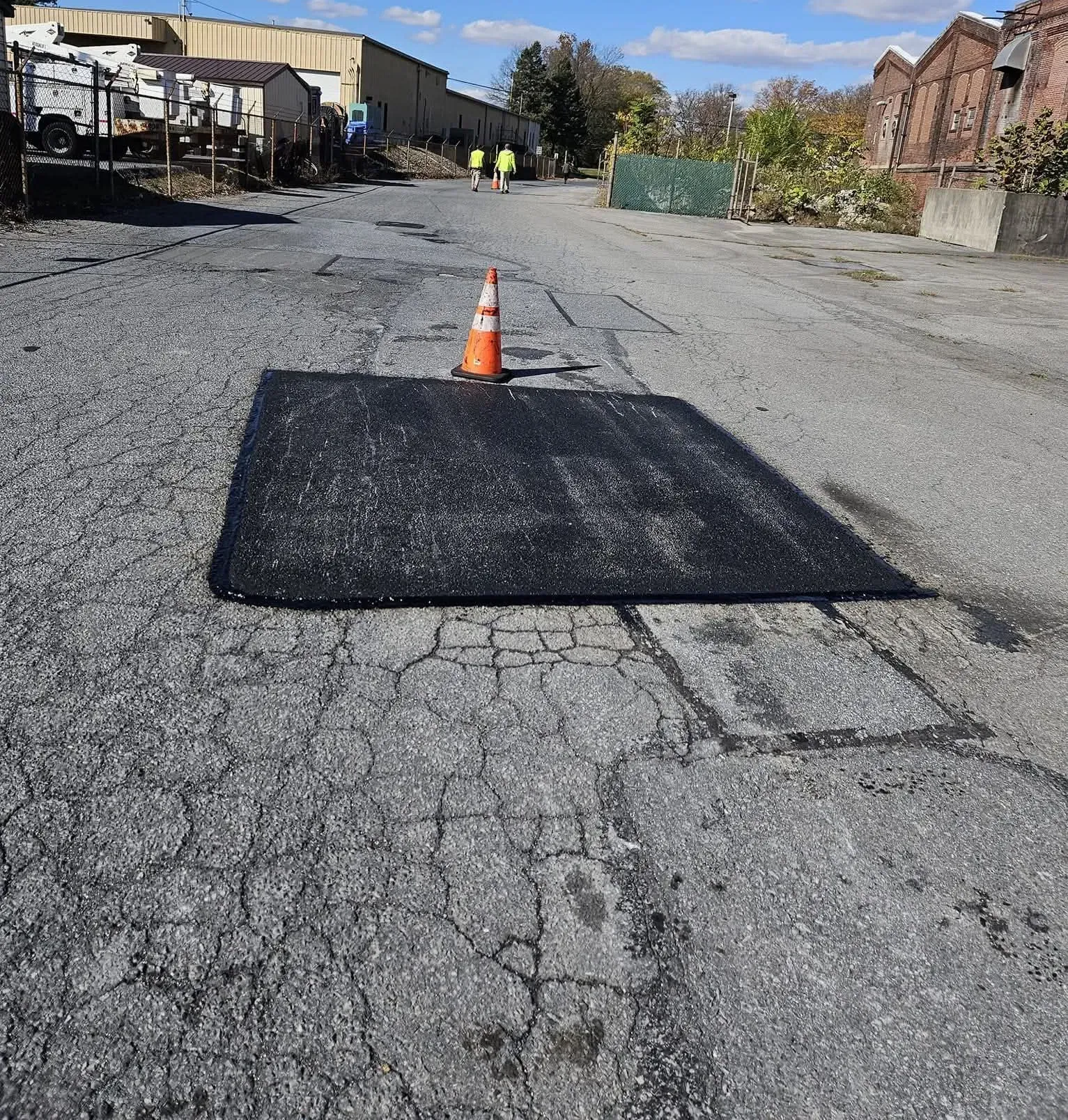 A black asphalt patch on a worn, cracked road, marked by a single orange traffic cone in an industrial area.