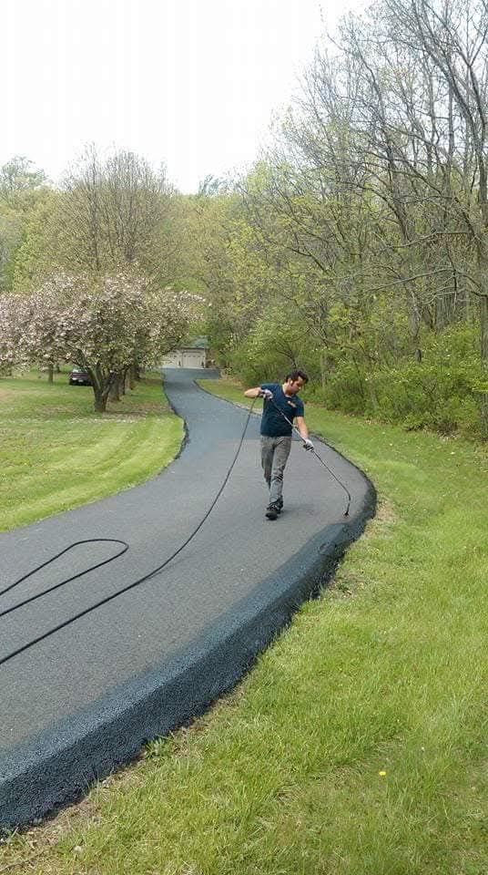 A person applying dark sealant to the edge of a curved asphalt driveway bordered by green grass and trees.