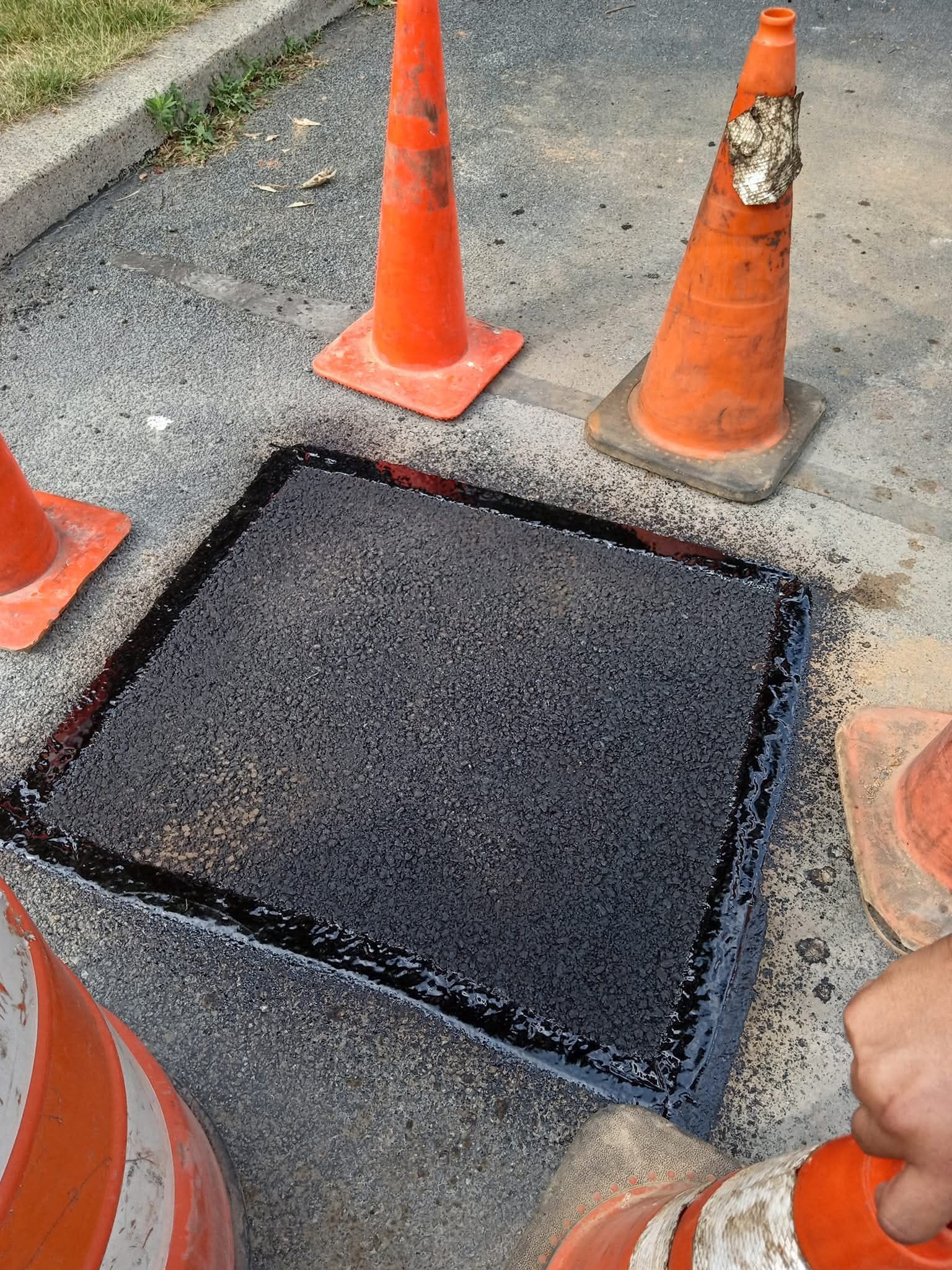 Asphalt road repair around a square utility access cover, with orange traffic cones.