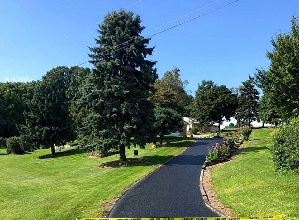 Newly paved asphalt driveway through a grassy yard, surrounded by trees under a blue sky.