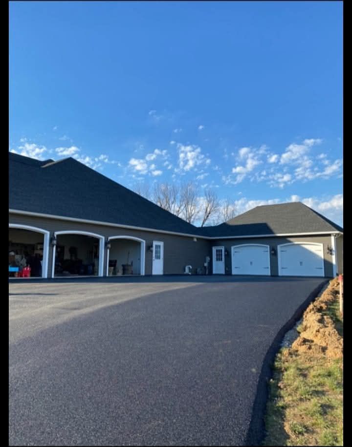 Dark asphalt driveway leads to a gray house with three garage doors under a blue sky.