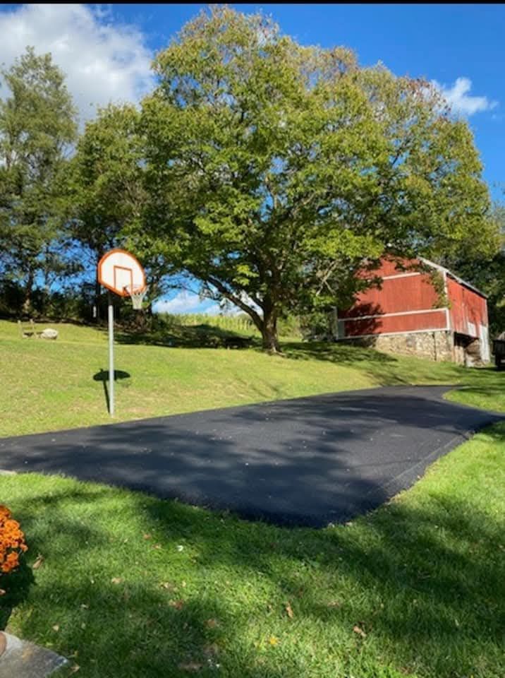 Basketball hoop on a paved court, large tree, red barn, green lawn under a blue sky.