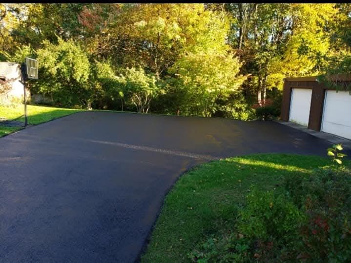 Newly paved asphalt driveway with a basketball hoop and garage, surrounded by grass and trees.