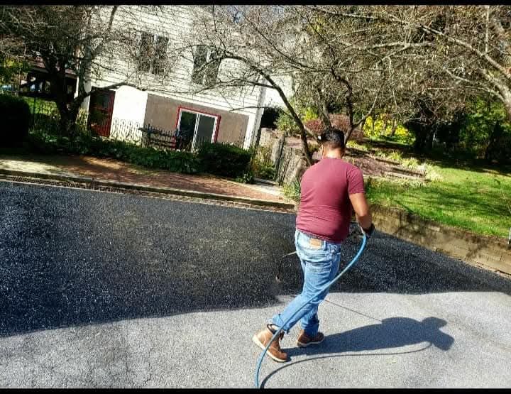 Man in a burgundy shirt and jeans washes a driveway with a hose on a sunny day.