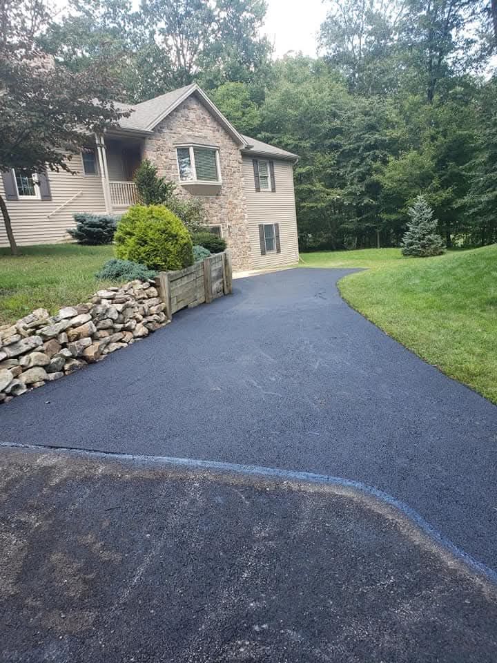 Dark paved driveway leading to a two-story house with stone facade, surrounded by green grass and trees.