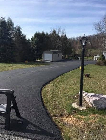 Newly paved black asphalt driveway curves through a grassy yard toward a white building and trees under a blue sky.