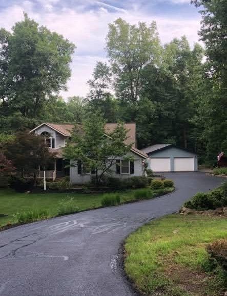 House with light blue siding, a two-car garage, and a winding asphalt driveway surrounded by greenery.