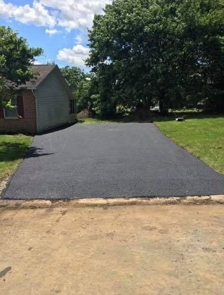 Newly paved black asphalt driveway next to a grassy lawn and a house under a blue sky.