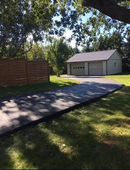 Asphalt driveway leads to a two-car garage with light-colored siding, under a shady tree, near a wooden fence.