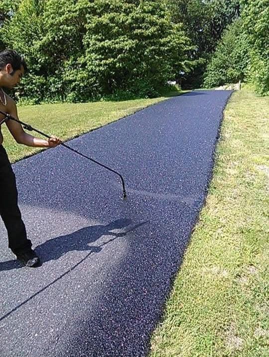Person spraying a newly paved asphalt path with a black coating, surrounded by green grass and trees.