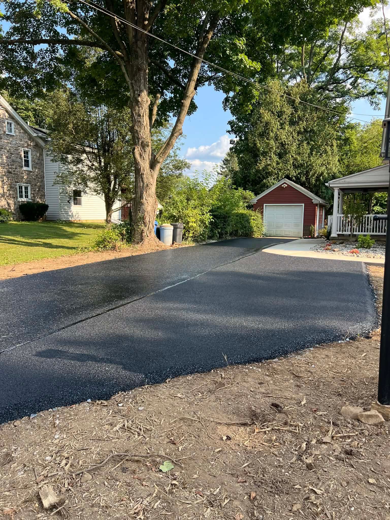 Newly paved asphalt driveway leading toward a red garage, beside a grassy lawn and a stone house.