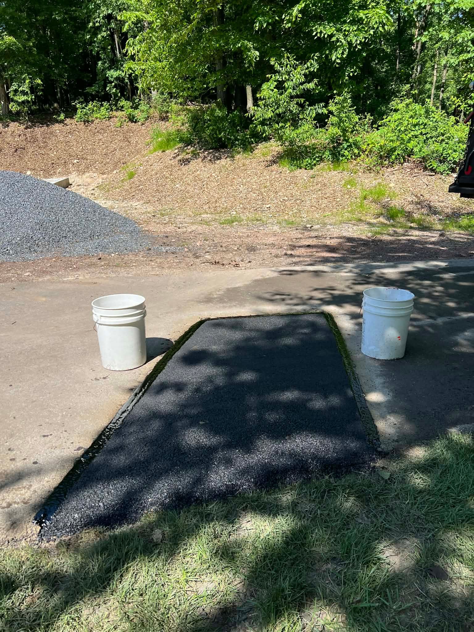 Black rubber playground surface being installed on concrete, with gravel and buckets nearby.