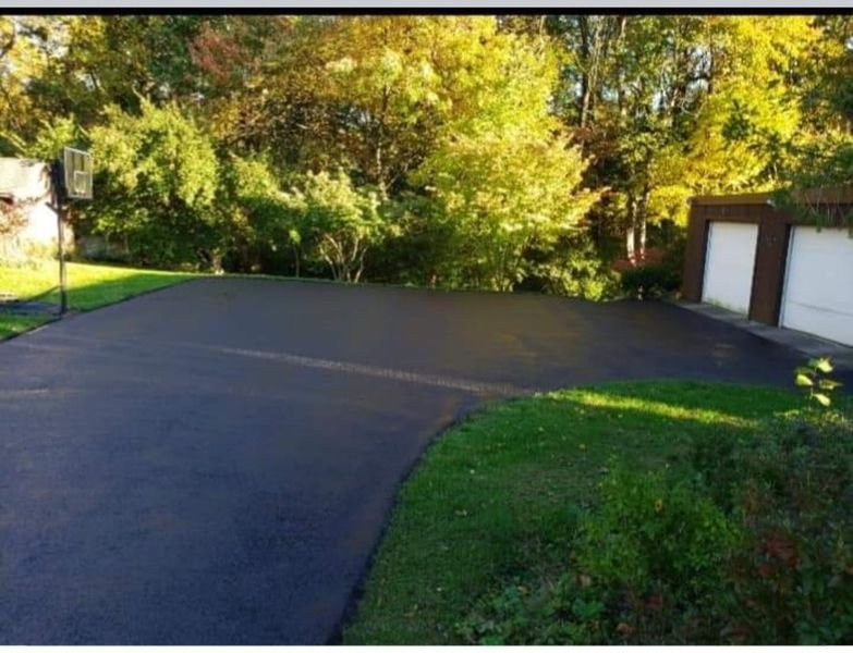Newly paved black asphalt driveway next to green lawn, with trees in the background and a garage.