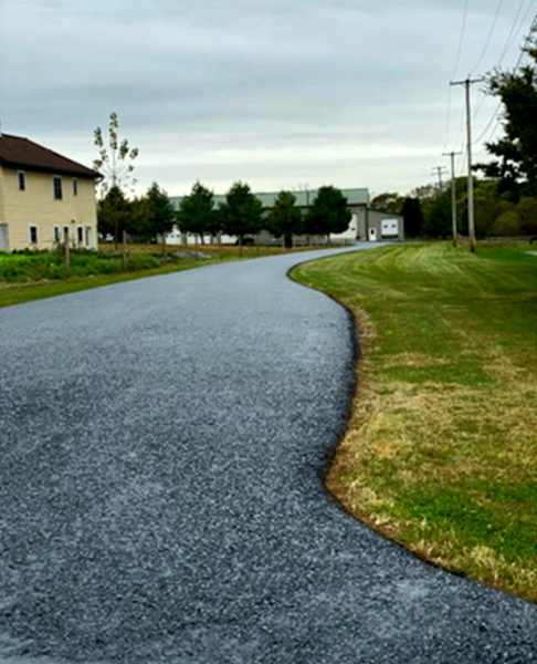 Paved road curves through a grassy area, buildings and trees in the background under a cloudy sky.