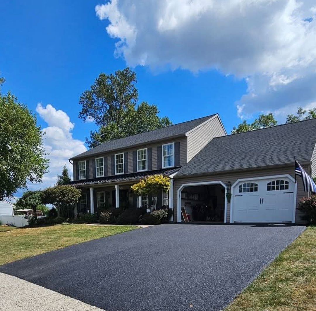 A two-story gray house with a white garage door, an asphalt driveway, and a large tree against a blue, cloudy sky.