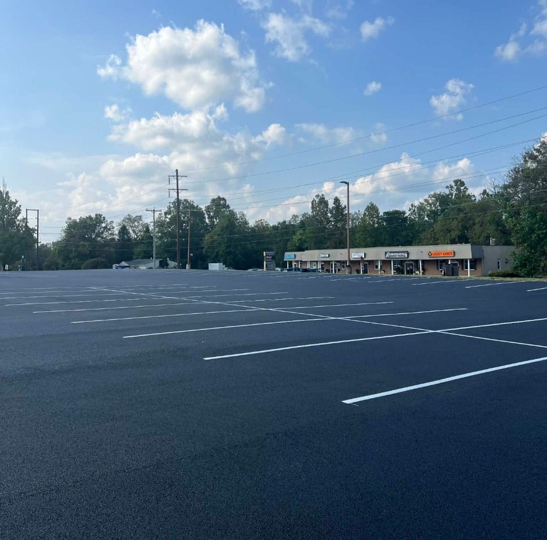 Newly paved, dark asphalt parking lot with white painted lines under a bright, sunny blue sky with a strip mall beyond.
